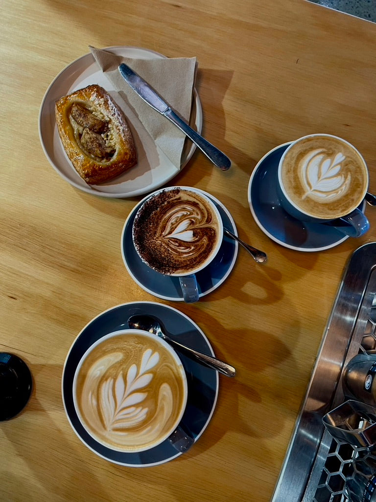 Three coffees with latte art and a pastry on a timber café table
