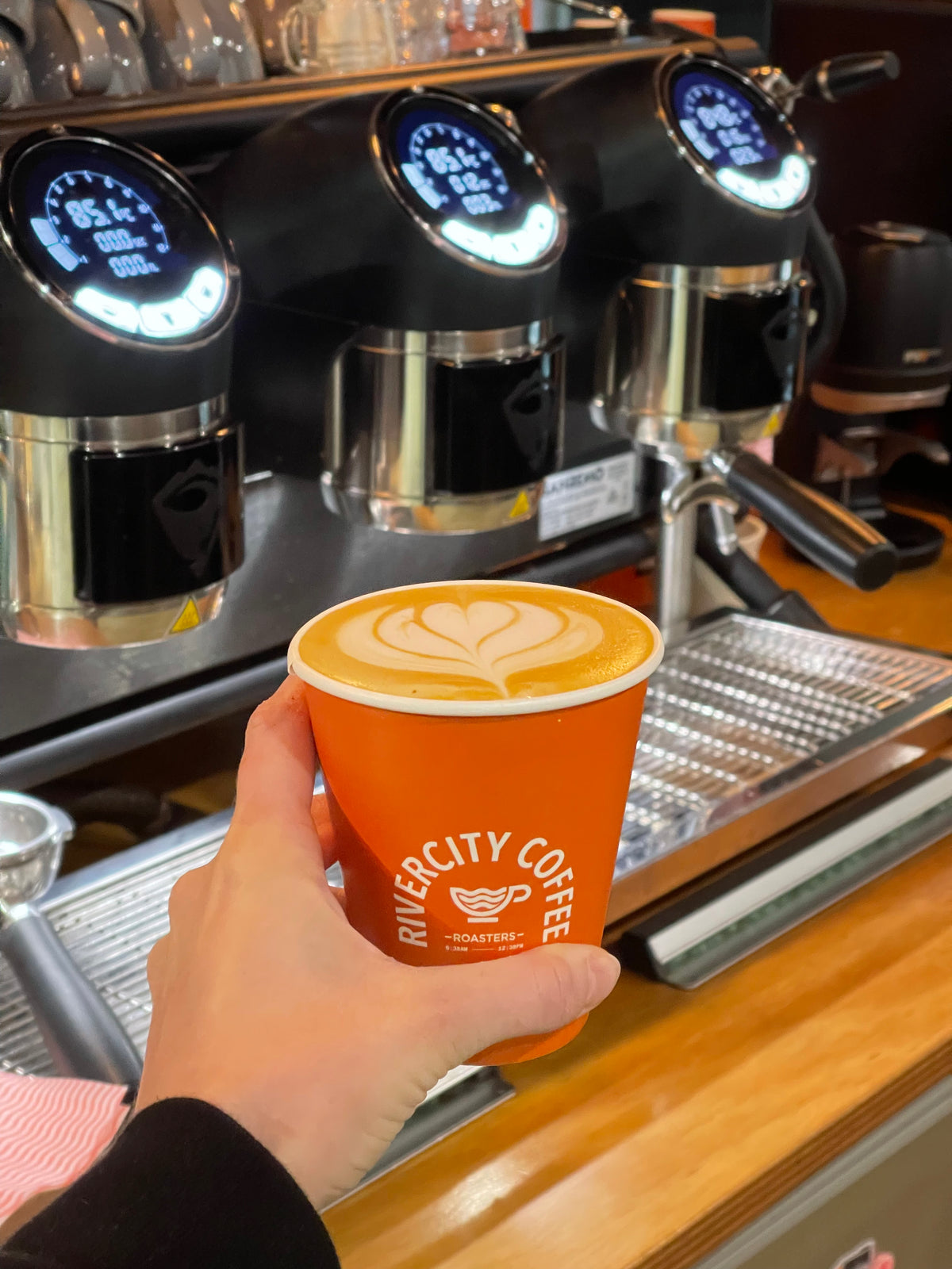 Hand holding an orange River City Coffee Roasters takeaway cup with latte art at the espresso machine